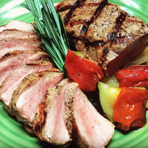 Grilled pork tenderloin with char marks, sliced and garnished with rosemary, peppers, and potatoes on a green plate.