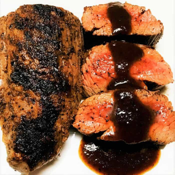 Pepper-crusted Teres Major steak sliced and topped with barbecue sauce on a white plate