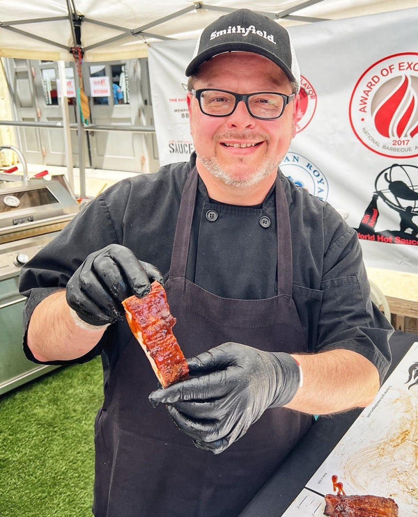 Smiling chef in black apron holding sauced barbecue ribs at a competition booth