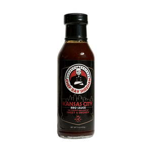 A bottle of Chef Ray Sheehan's Kansas City BBQ Sauce on a transparent background. The bottle is made of dark glass showing the dark brown sauce inside, with a black cap and a black label featuring the brand logo and product name in red and white text.